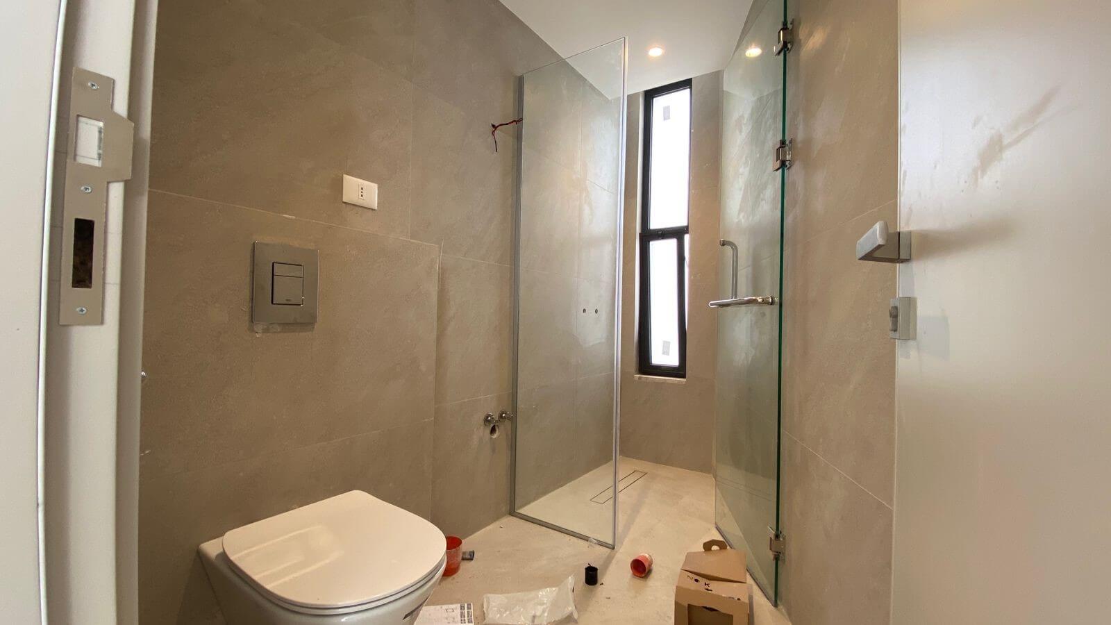 Unfinished modern bathroom featuring a wall-mounted toilet, beige tile walls, and a glass shower enclosure with a tall narrow window. Construction items and packaging debris are on the floor.