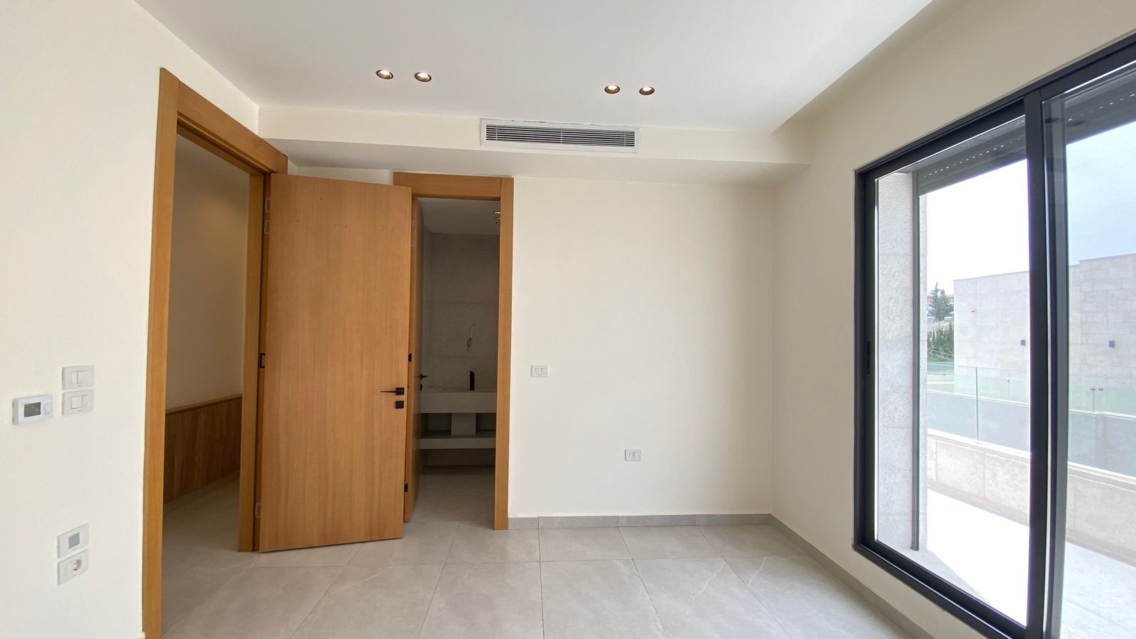 037e82d8-d225-4929-981b-5927b8c30452 Empty modern room with large window and balcony, light walls, tiled floor, and open wooden doors to a hallway.