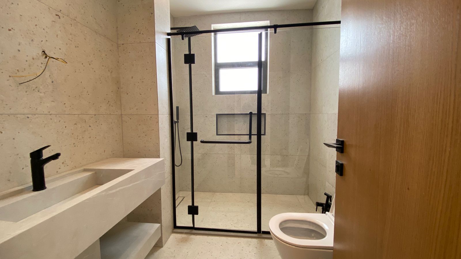 0413242a-9699-4e56-acbd-ac49740d368d Contemporary bathroom with beige tiled walls, glass walk-in shower with black metal frame, a white sink, and a toilet visible to the right.