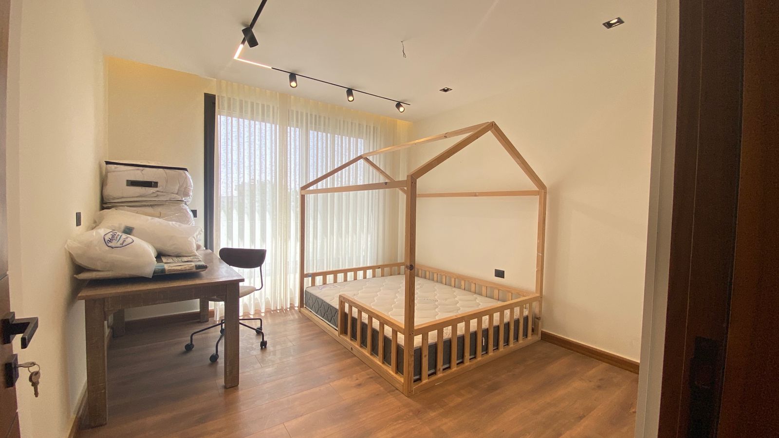 048dd3e0-b8ef-4c53-8c5d-c897ef5005f7 Bedroom with a wooden house-frame bed and a mattress, placed on light wood flooring; there is a desk and a black rolling chair to the left, and large windows with sheer white curtains letting in daylight.