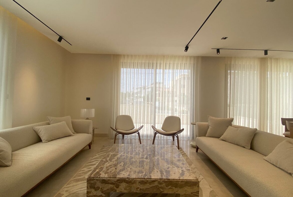 Neutral, sunlit living room with two long beige sofas facing a marble coffee table and two white lounge chairs by large windows with sheer curtains