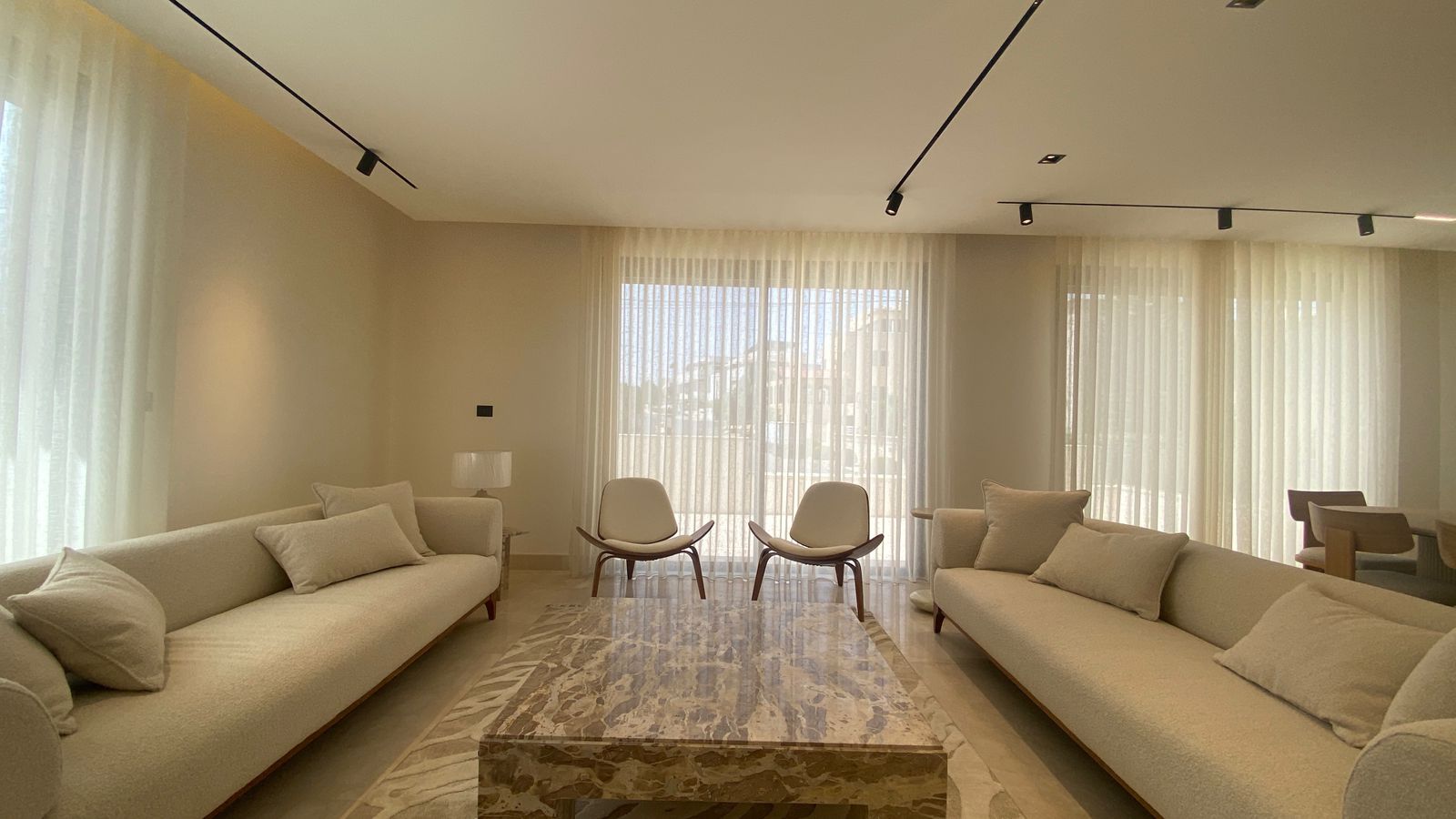 04cda819-a893-42d2-a450-c728717b8c46 Neutral, sunlit living room with two long beige sofas facing a marble coffee table and two white lounge chairs by large windows with sheer curtains