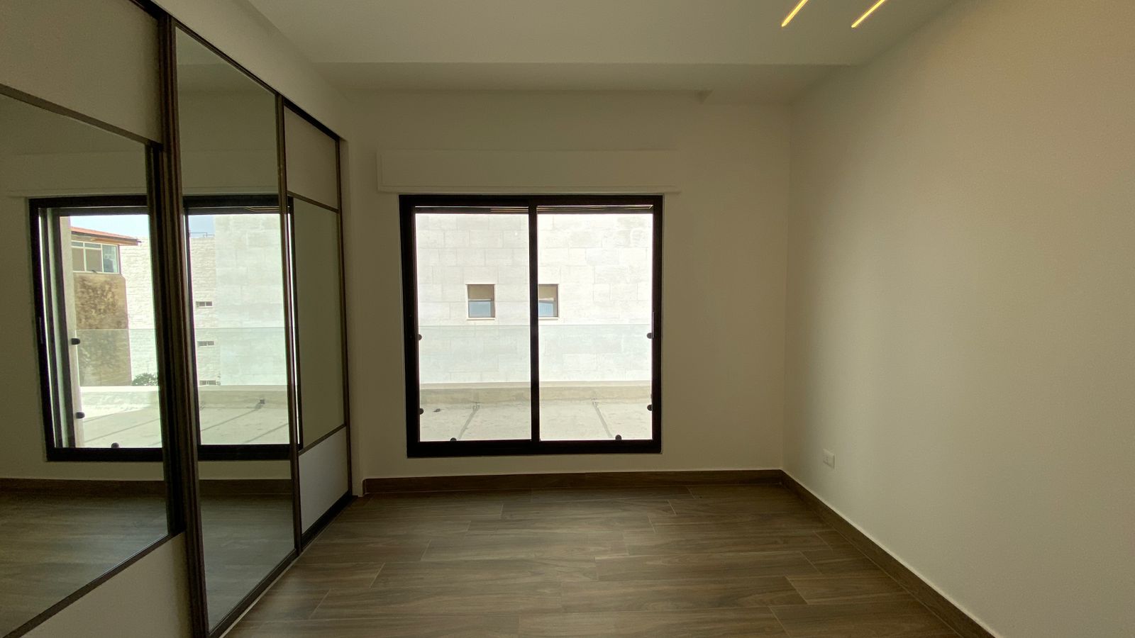 05092f0d-a366-4447-b7d4-85049d742031 Empty, unfurnished room with a large glass door/window opening to a light outdoor space and a mirrored wardrobe on the left.