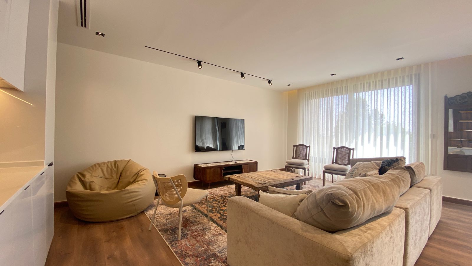 051aa581-1809-4918-a870-5c230e56edcc Spacious living room with a beige sectional sofa facing a wall-mounted TV, a wooden coffee table, and a patterned rug underfoot.