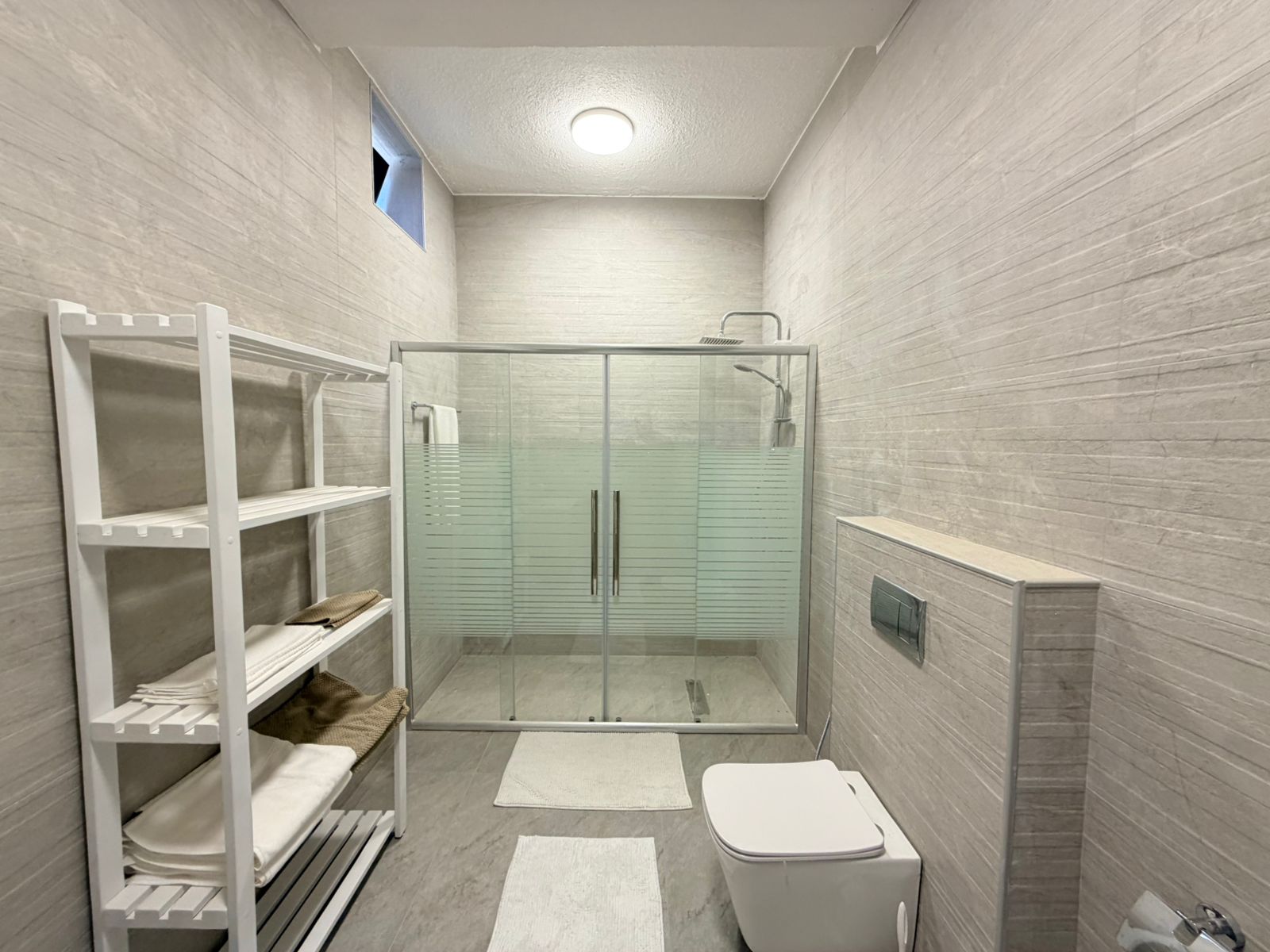 0596298f-9f01-4983-986d-1922c3849f96 Modern bathroom with a glass-enclosed shower, white toilet, and open white shelving holding towels and woven items. Light gray tiled walls and floor create a clean, minimalist look.