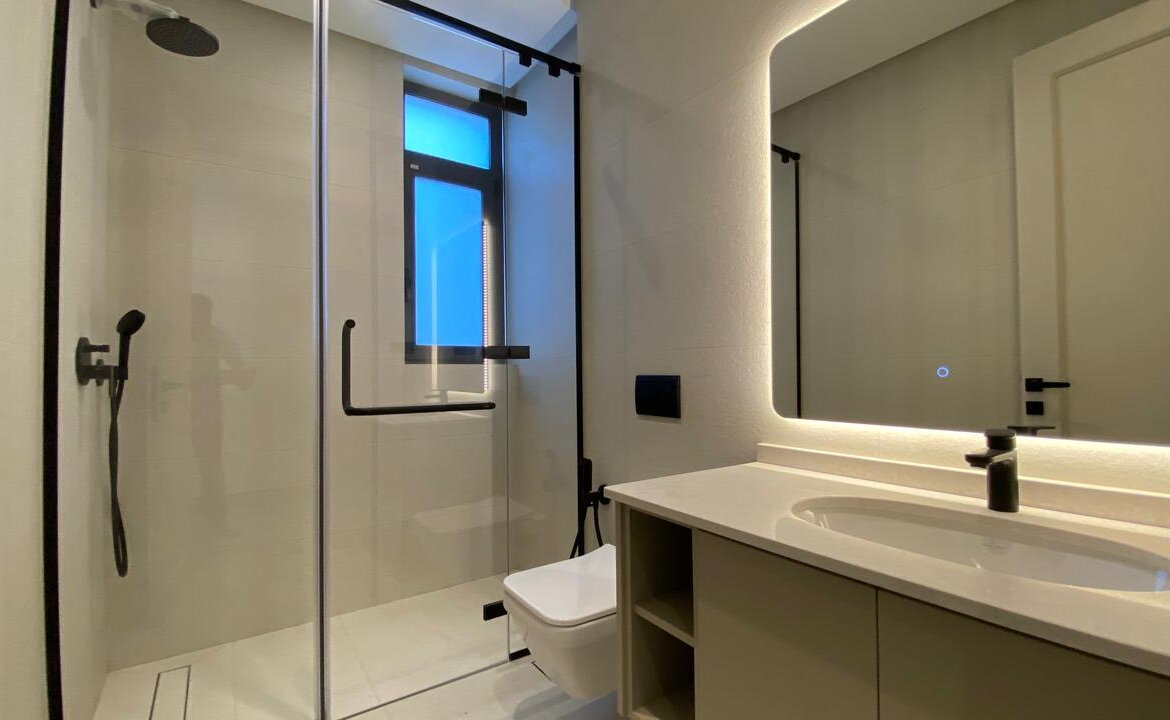 Modern bathroom with glass shower enclosure, wall-mounted toilet, and a white countertop sink under a backlit mirror