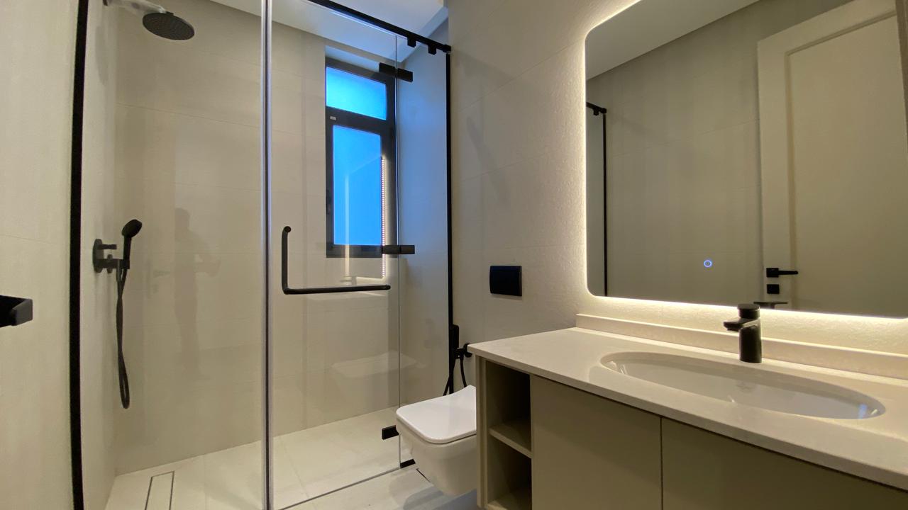 Modern bathroom with glass shower enclosure, wall-mounted toilet, and a white countertop sink under a backlit mirror