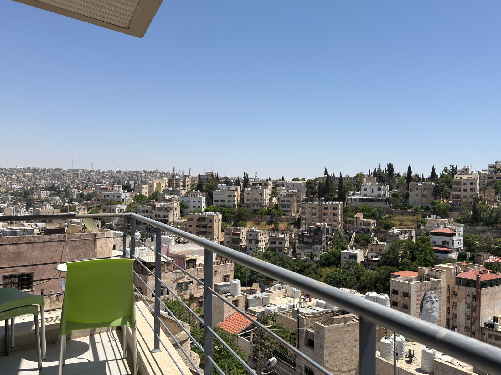 066837c8-f74b-4717-9009-52fab9a2962a City skyline viewed from a balcony with a metal railing, beige apartment blocks, and a clear blue sky above.