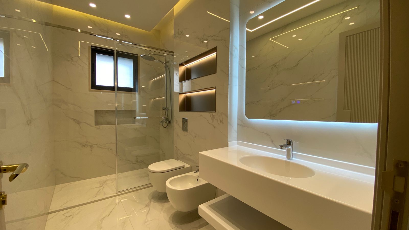 06b5096a-302d-4d08-a7e5-98b9a57721db Luxurious marble bathroom with glass shower enclosure, wall-mounted sink, and a bidet beside a wall toilet.
