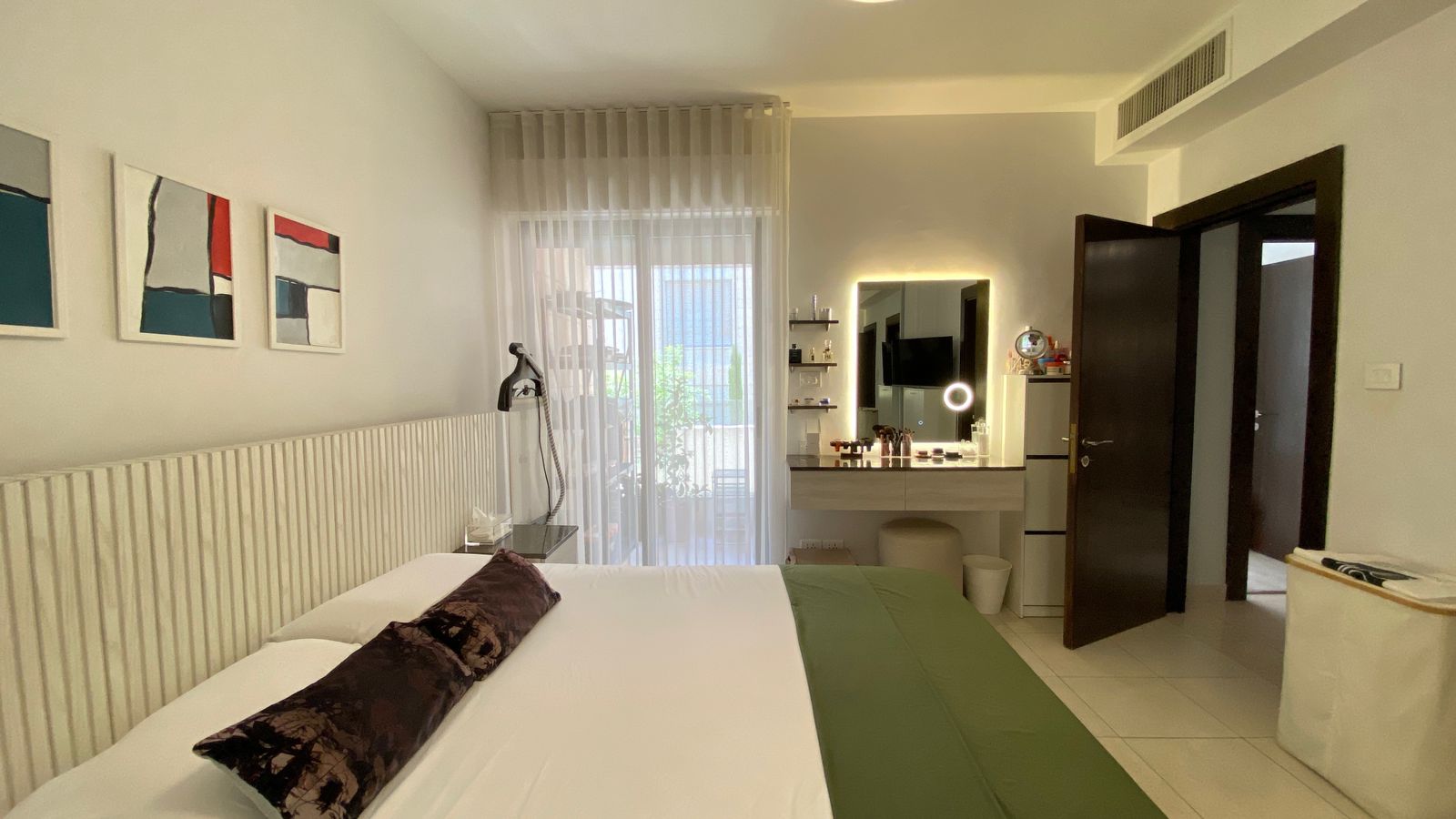 07069fcb-446a-4e32-84ab-3a802c08252d Hotel room with a large bed, sheer curtains, and a makeup vanity with ring light opposite the foot of the bed.