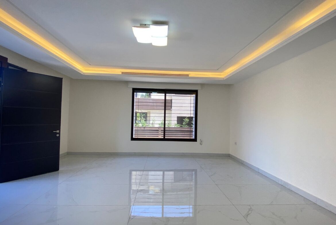 Empty modern living room with glossy white tile floor, warm LED cove lighting, an open dark door to the outdoors, and a large window overlooking a courtyard.