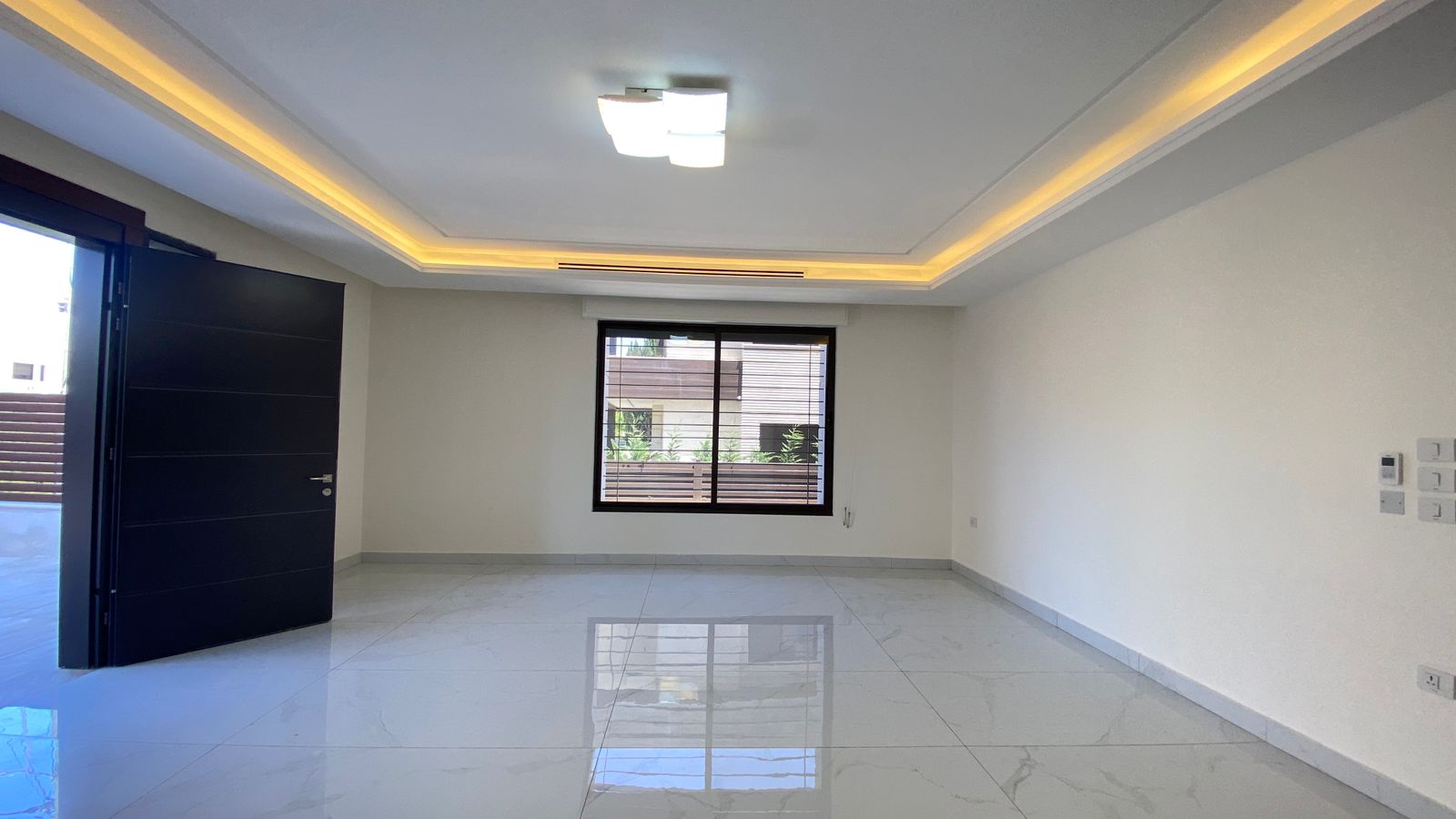 0734ed7f-0f12-4760-9234-339ae923c247 Empty modern living room with glossy white tile floor, warm LED cove lighting, an open dark door to the outdoors, and a large window overlooking a courtyard.