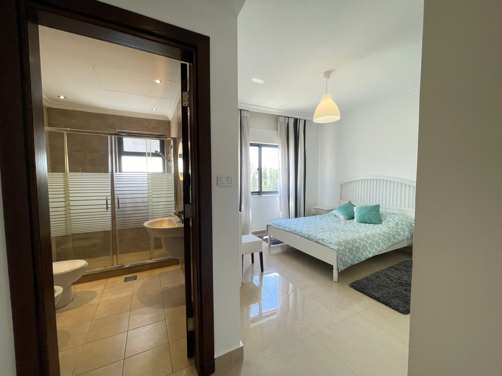 0744aad4-44f3-4db1-aac8-5791140427f9 Bedroom with white metal bed, mint-green bedding, and two teal cushions; window with sheer curtains nearby, pendant light, rug on light tiled floor, adjacent bathroom visible through doorway with glass shower enclosure.