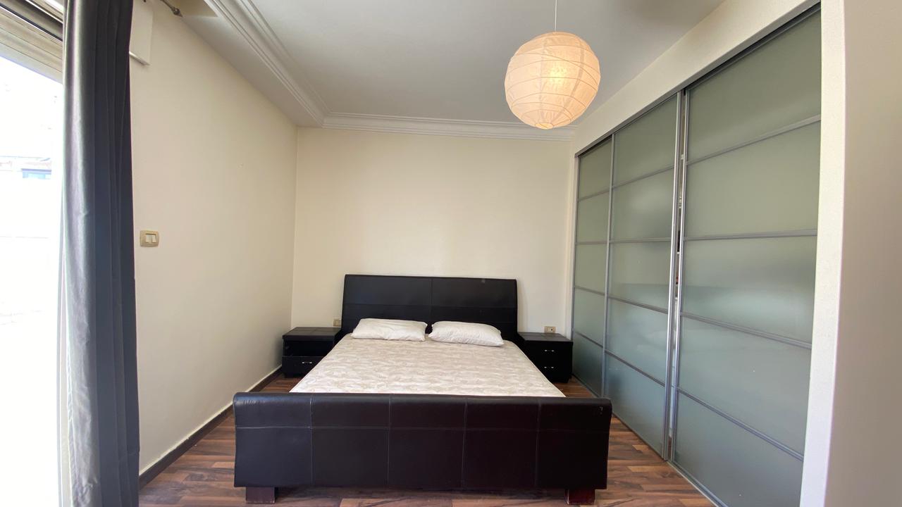 077d5f21-5671-47ce-b2ea-66f4d2abe0de Bedroom with a dark bed frame, two matching nightstands, and a pale mattress ready for bedding; frosted-glass sliding wardrobe on the right; round paper lantern ceiling light above.