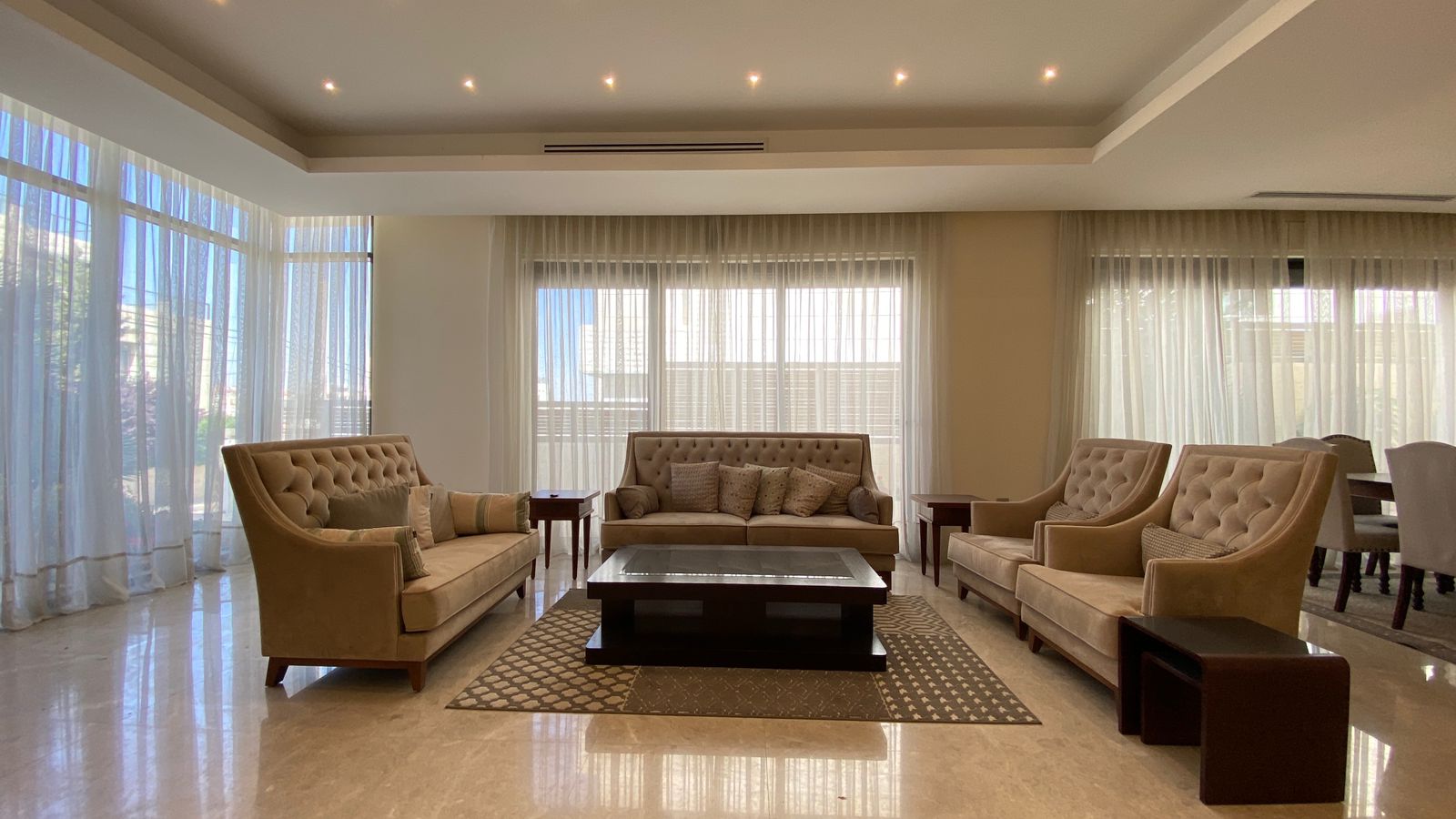07c6d97e-bf96-4fe3-be07-15b354885424 Spacious beige living room with tufted sofas, matching chairs, and a dark wood coffee table on a patterned rug.