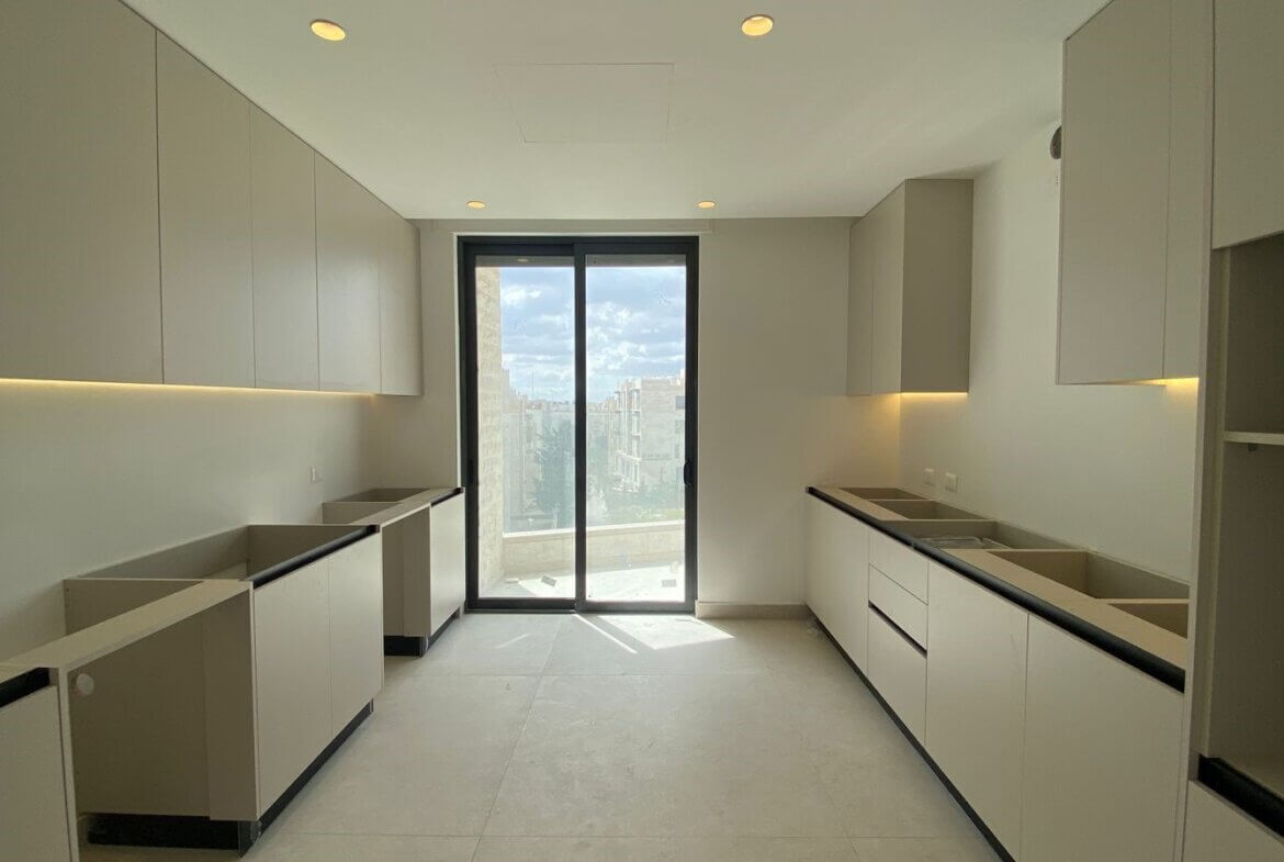 07cd23f6-f03e-4a05-b534-a3c7c6622ebc Modern, minimalist kitchen (galley layout) with light-gray cabinets on both sides, under-cabinet lighting, and a glass door to a balcony overlooking the city skyline.