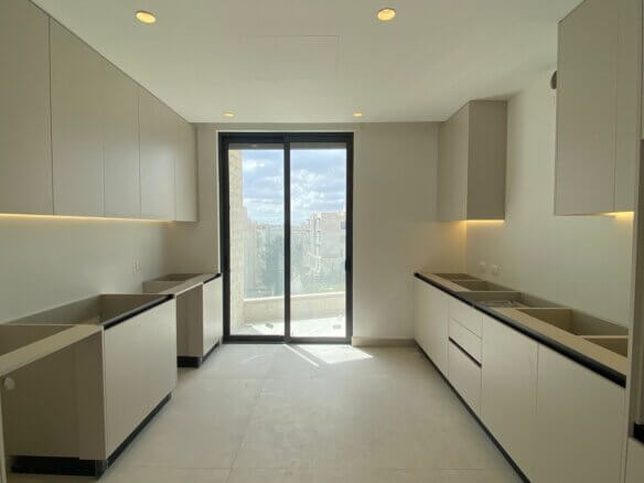 Modern, minimalist kitchen (galley layout) with light-gray cabinets on both sides, under-cabinet lighting, and a glass door to a balcony overlooking the city skyline.