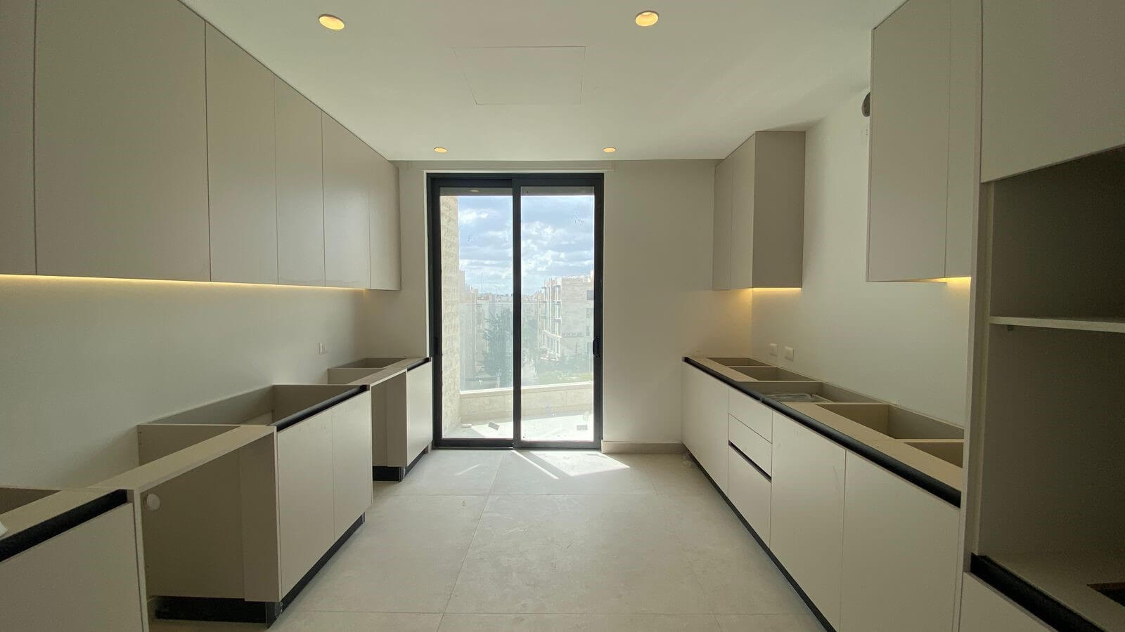 07cd23f6-f03e-4a05-b534-a3c7c6622ebc Modern, minimalist kitchen (galley layout) with light-gray cabinets on both sides, under-cabinet lighting, and a glass door to a balcony overlooking the city skyline.