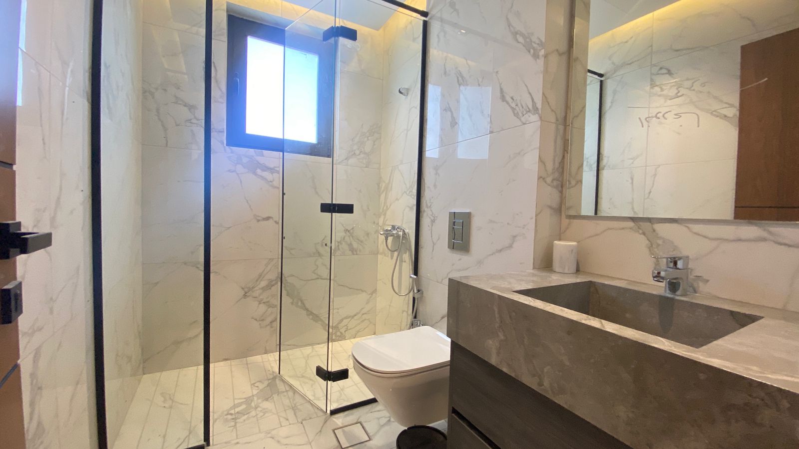 07fa7ac1-6c45-488c-a3cf-3a868558ee9c Modern marble bathroom featuring a glass-enclosed shower with black metal frame, wall-mounted toilet, and a gray stone vanity with sink.