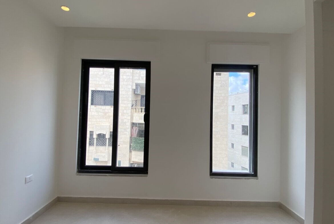 0a0ca71d-2b2a-4ed0-8c9d-8f3660b25c5c -empty room with white walls, two tall black-framed windows, and a light gray tiled floor; view of a beige building outside; recessed ceiling lights visible