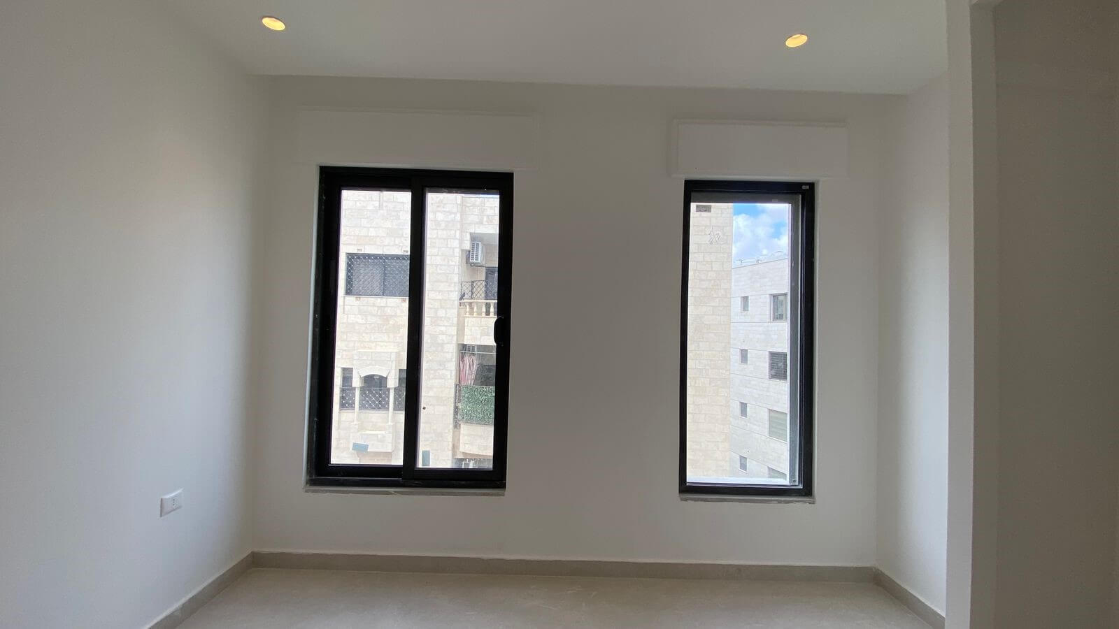 0a0ca71d-2b2a-4ed0-8c9d-8f3660b25c5c -empty room with white walls, two tall black-framed windows, and a light gray tiled floor; view of a beige building outside; recessed ceiling lights visible