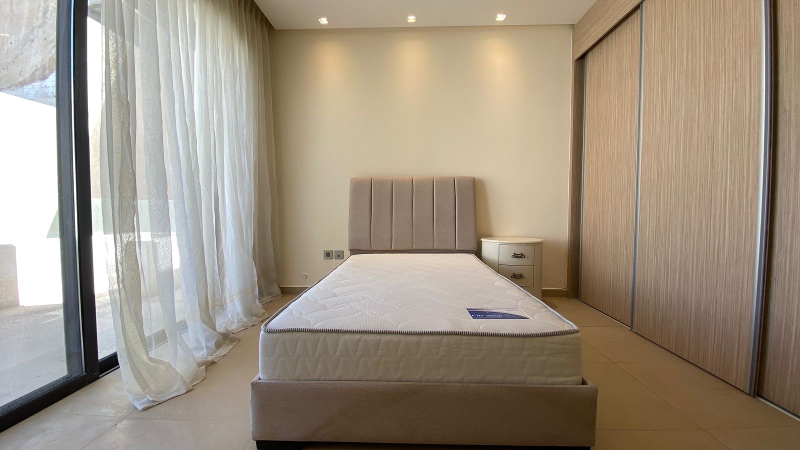0ac05b75-7a5a-4efc-99b0-6210842d56ec Modern bedroom with a low beige bed, tall upholstered headboard, and a white nightstand in a bright, minimalist space.