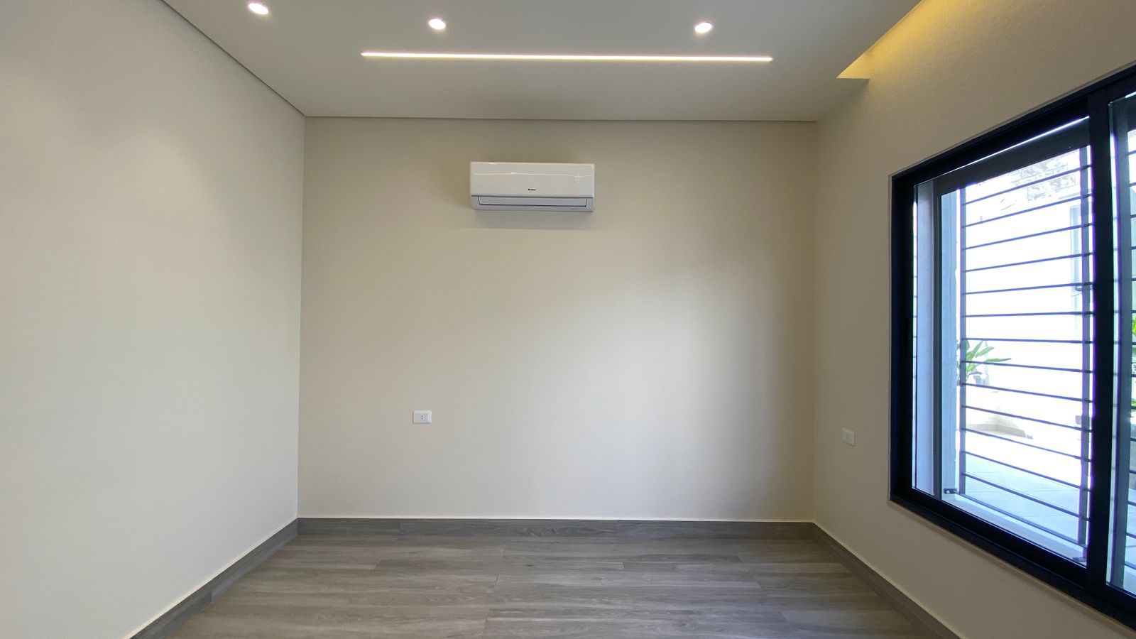 0ac2050c-7e83-4d75-8bb1-da5b33960692 Empty modern room with gray wood-like flooring, white walls, a wall-mounted air conditioner, recessed ceiling lights, and a large window with metal bars on the right.