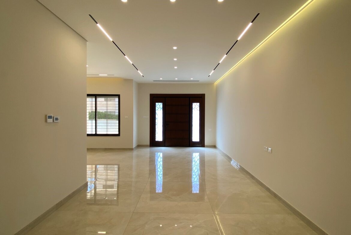 Empty living room with glossy tiled floor, beige walls, and linear ceiling lights leading to a dark wooden front door with side panels.