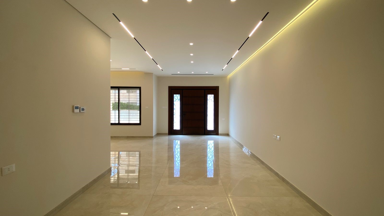 0b0ee7aa-55ad-4841-bd41-8563c16a97e7 Empty living room with glossy tiled floor, beige walls, and linear ceiling lights leading to a dark wooden front door with side panels.