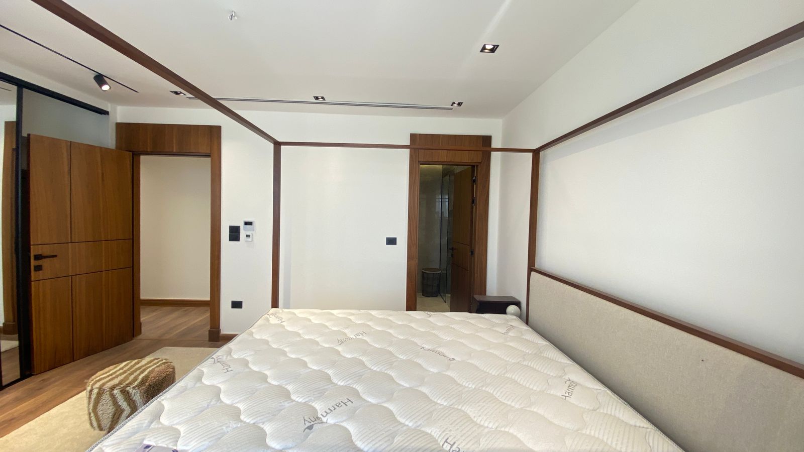 0be11878-7c4f-4738-8e36-1e9349c41c16 Bedroom with a large bed in the foreground, beige headboard, and wood trim; a tall wooden wardrobe on the left and an open door to an en-suite bathroom nearby.