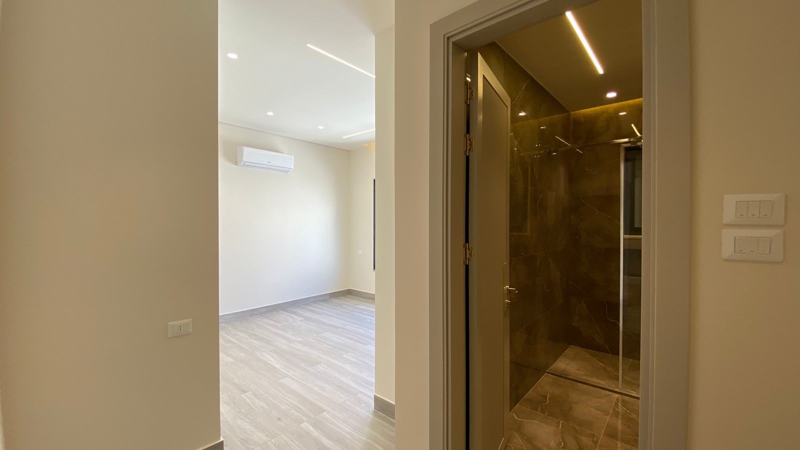 0be828ae-589e-4e51-bd98-e8c7399ab311 Empty modern room with light wood flooring and a wall-mounted AC, seen from a doorway, leading to a marble-tiled bathroom with a glass door.