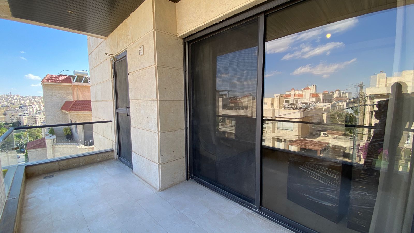 0bfa7152-321f-4334-84f5-2d3c1195f7d1 Balcony with glass railing overlooking a cityscape, beige stone walls, and a sliding glass door/reflection on the right side
