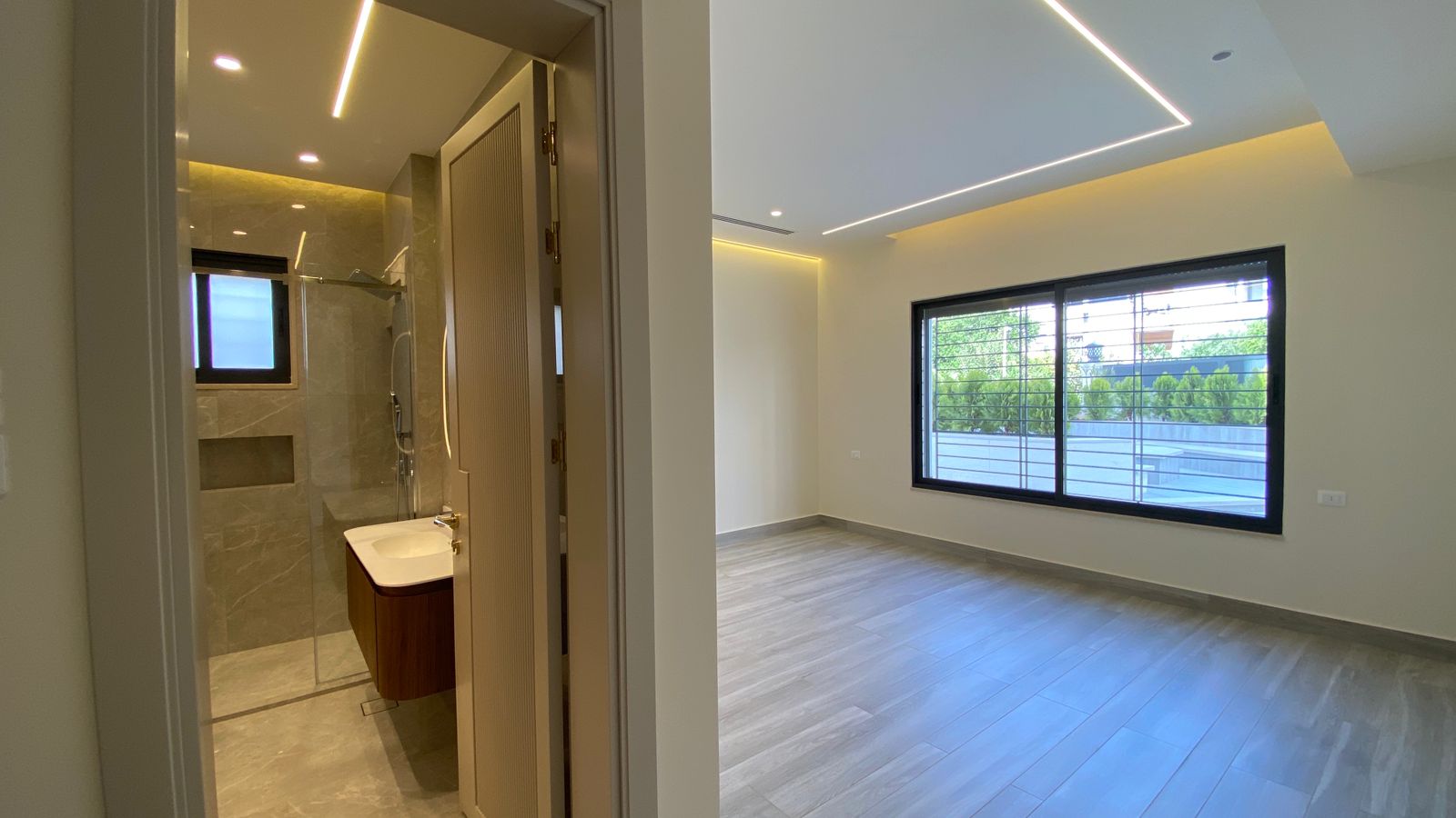 0cbb42e9-2cdd-4a8e-a7b1-066b85f702df Empty modern room with large window, gray wooden floor, and recessed ceiling lights; bathroom doorway on left shows glass shower and sink.