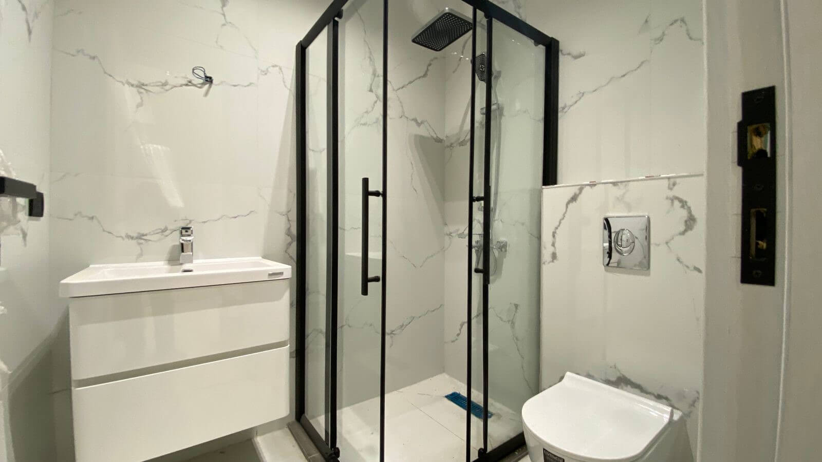 0cf80a43-1232-4801-ba1f-3e267a0d5b9f Modern bathroom with a glass-framed shower, white vanity, and wall-mounted toilet in a marble-tiled room.