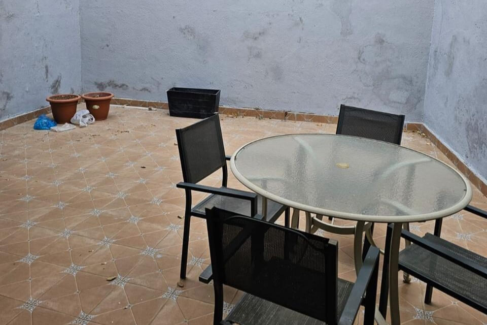 Outdoor patio with a round glass table and four black chairs on terracotta tiled floor; blue walls and potted plants in the corner.