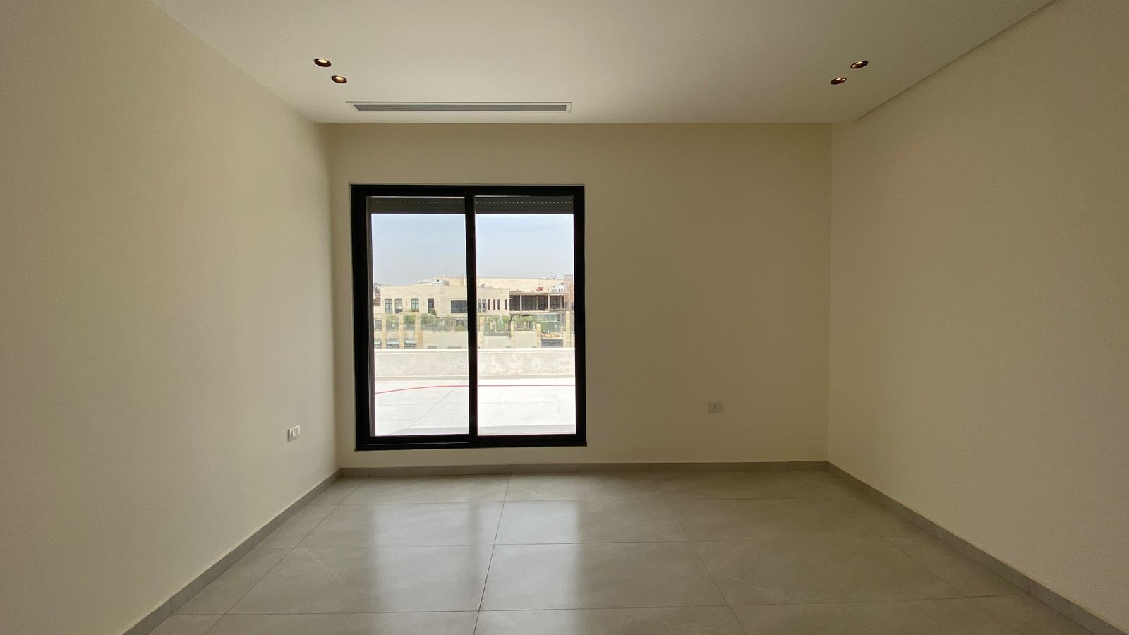 0e4c53c0-0bc2-4edb-a2d7-1ef3d16310bb Empty rectangular room with light beige walls, tiled gray floor, and a large black-framed window looking out to a building complex.