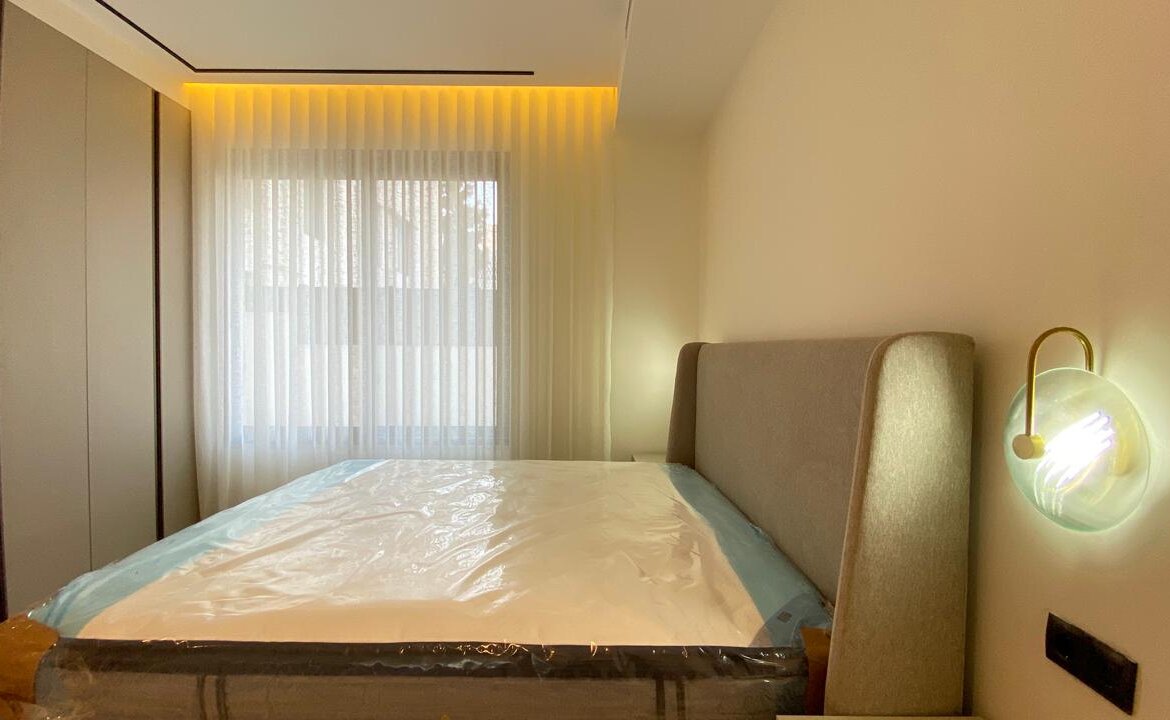 Modern hotel-style bedroom with a bed wrapped in plastic, beige headboard, and warm wall lamp beside the headboard