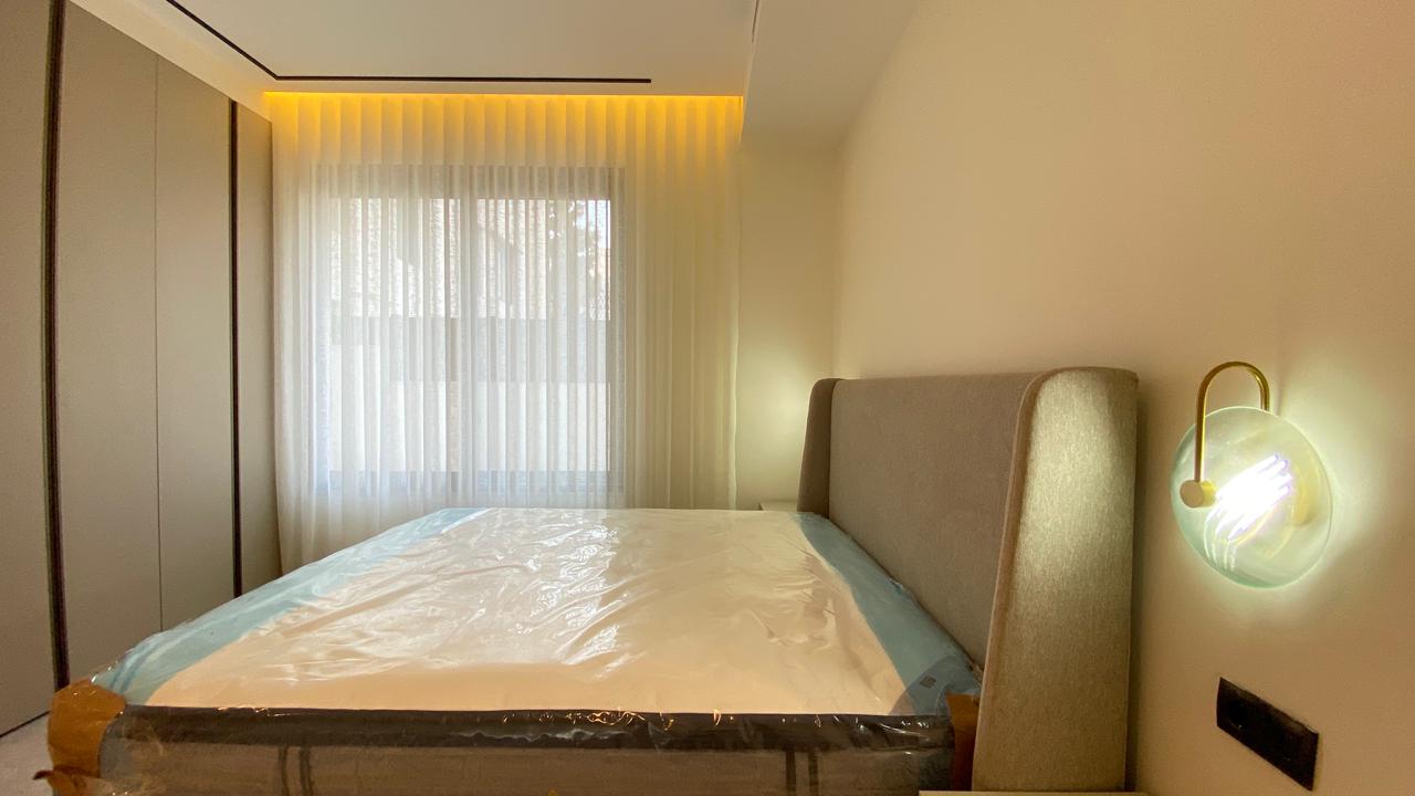 Modern hotel-style bedroom with a bed wrapped in plastic, beige headboard, and warm wall lamp beside the headboard