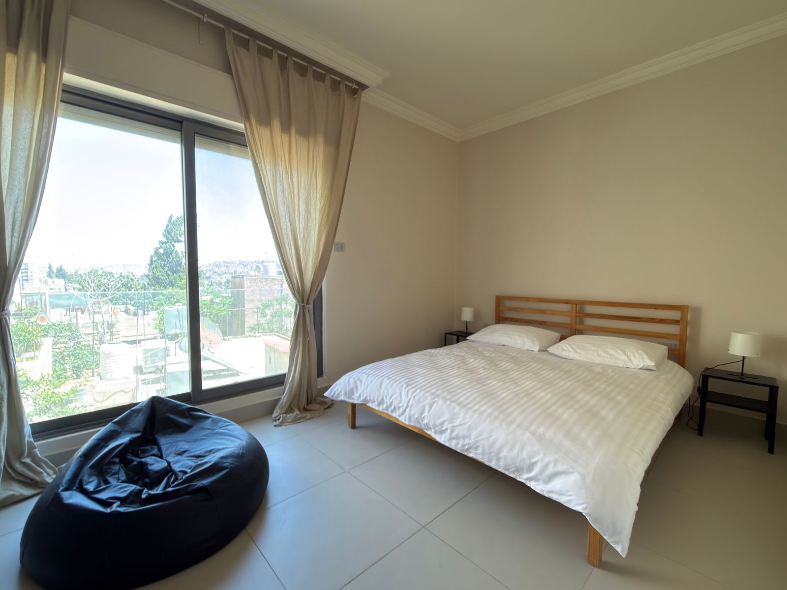 0ec8461f-4a0e-451f-9bbd-467632de4ff3 Bedroom with a wooden double bed, white striped comforter, two nightstands and lamps, near a large window with beige curtains.