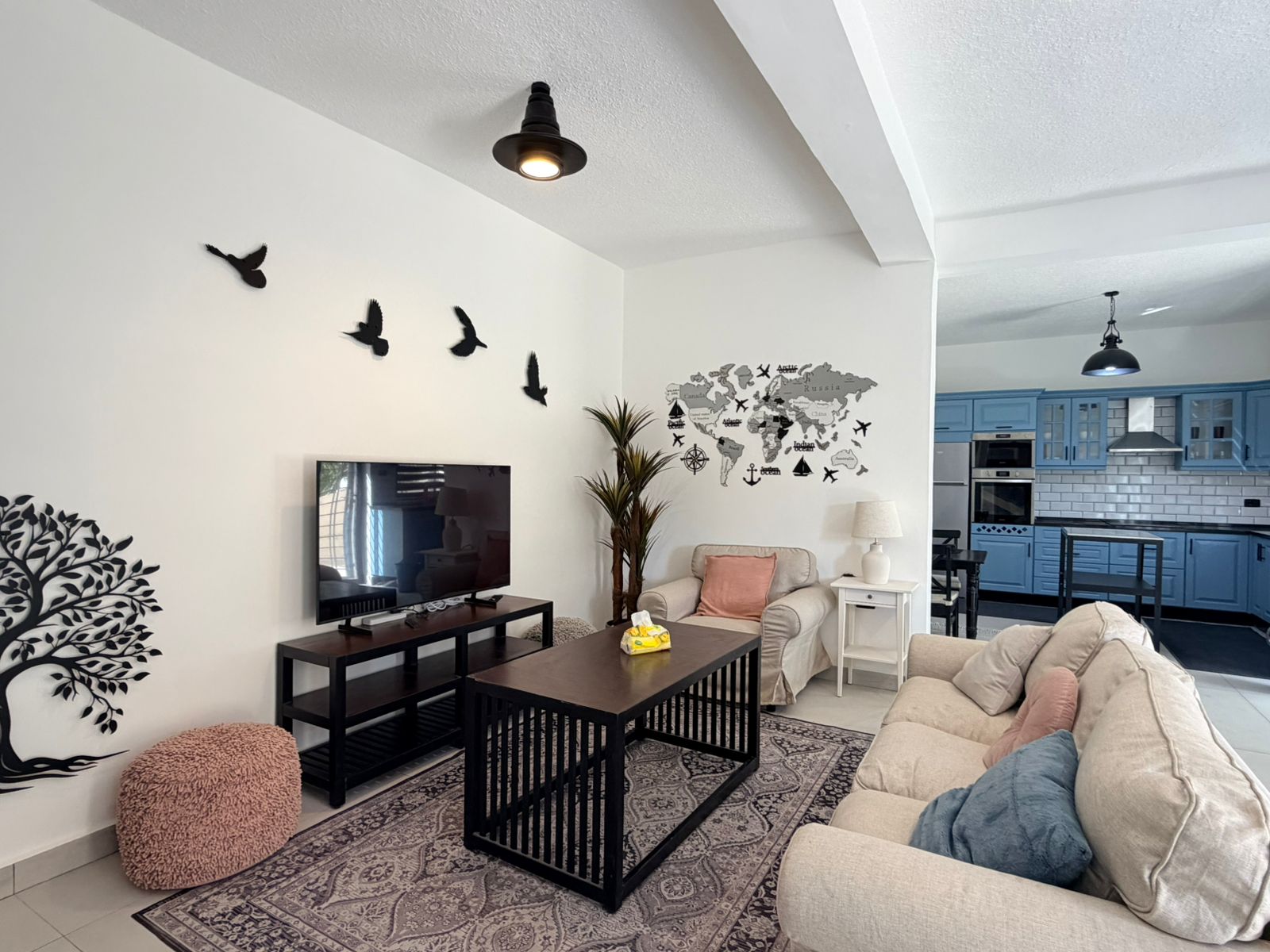 0eda42c9-e2ff-4937-a6b7-50f8f8de7546 Living room with beige sectional, coffee table, TV on a black stand, and wall decals of birds and a world map on the wall above.