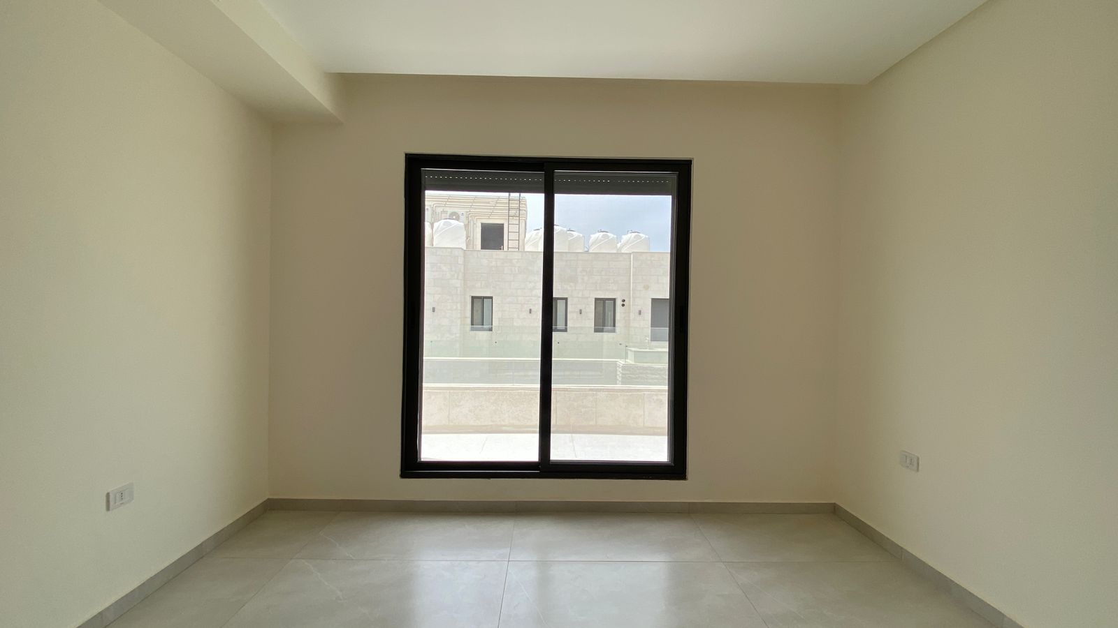 0ef6313d-c393-49f9-b169-1f74fedfeeb7 Empty room with beige walls and tiled floor, featuring a large black-framed sliding window facing a brick exterior building outside the window.