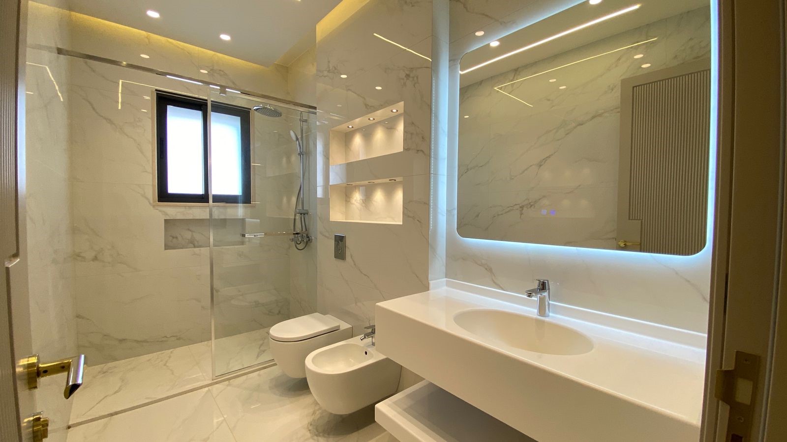 100e24d4-050a-4062-99bd-d2a09dea3e62 Modern marble bathroom with glass shower, wall-mounted toilet and bidet, and a white countertop sink with backlit mirror.