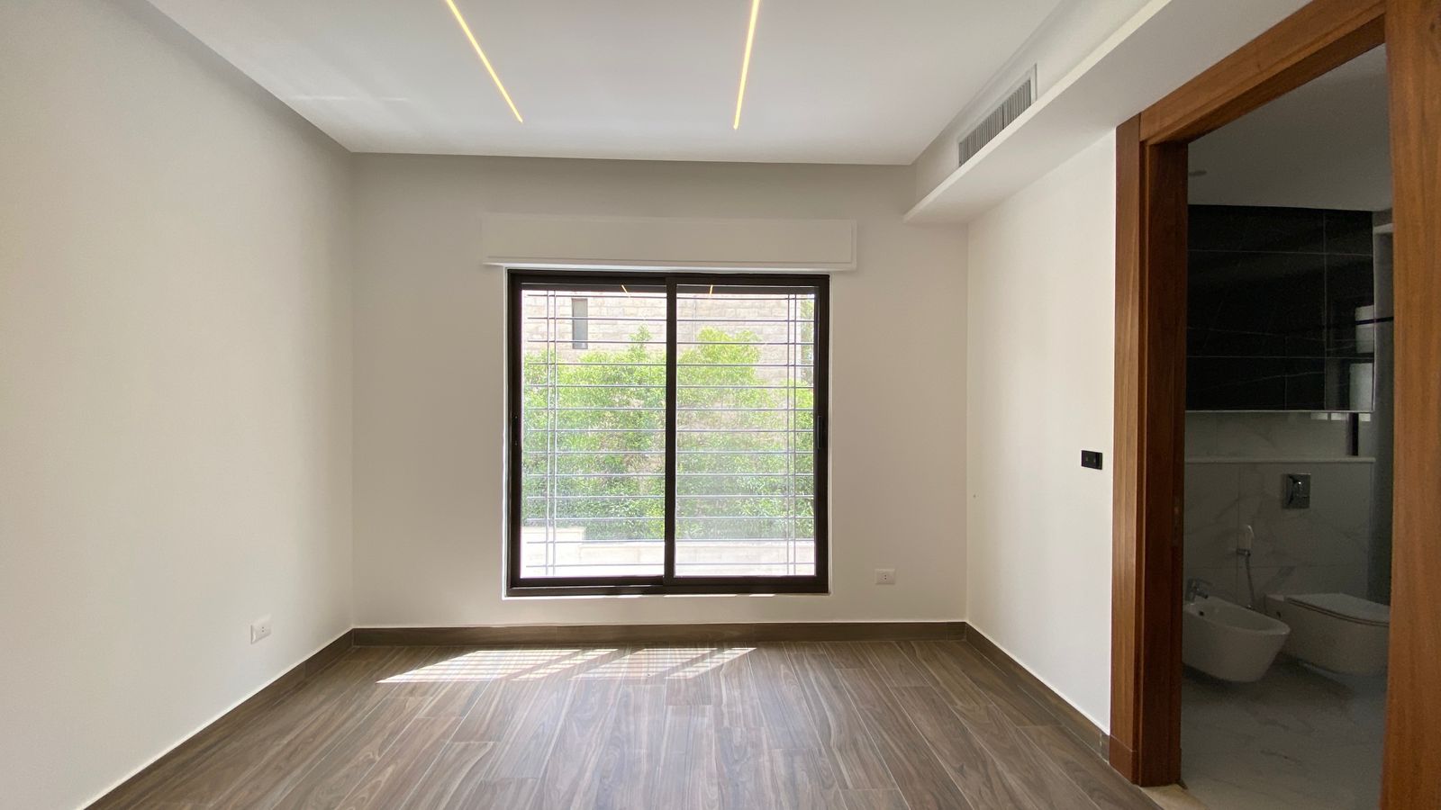10184c81-4df2-4608-a679-a034be07e15d Empty modern room with wood-look flooring, a barred window letting in light, and a doorway to a bathroom on the right.