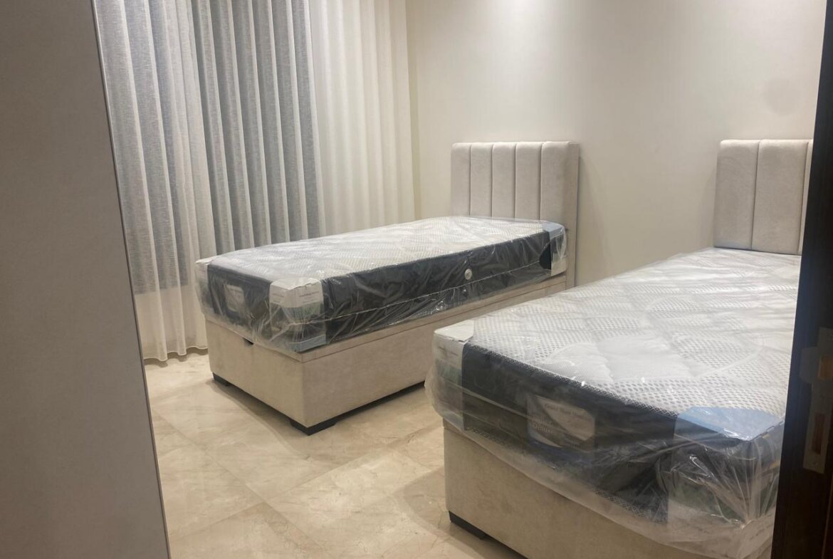 Bedroom showroom with two white upholstered twin beds still in plastic, beige headboards, and light marble floor.
