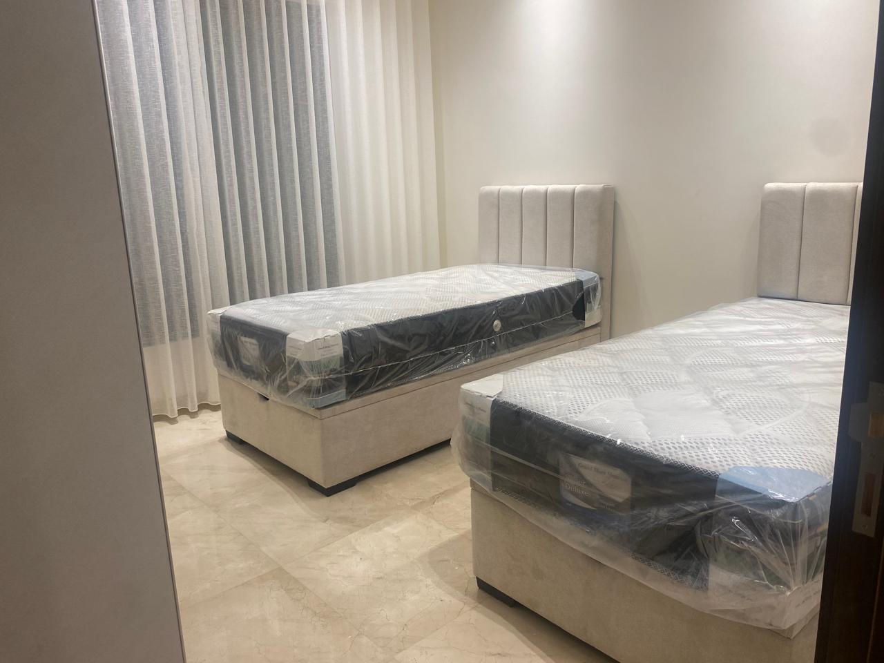 Bedroom showroom with two white upholstered twin beds still in plastic, beige headboards, and light marble floor.