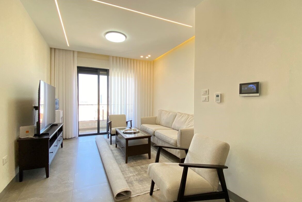 Bright modern living room with white sofa and matching armchairs, coffee table, TV on a dark console, and a rolled rug in front of the sofa; balcony doors with sheer curtains in the background.