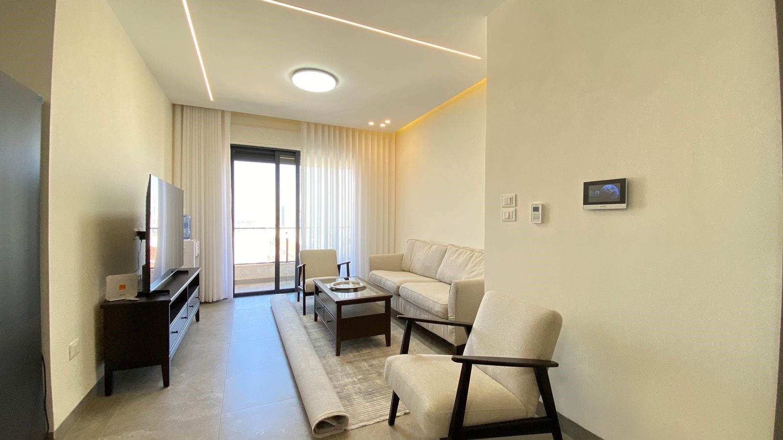 1258923b-f977-483b-91e5-594442870358 Bright modern living room with white sofa and matching armchairs, coffee table, TV on a dark console, and a rolled rug in front of the sofa; balcony doors with sheer curtains in the background.