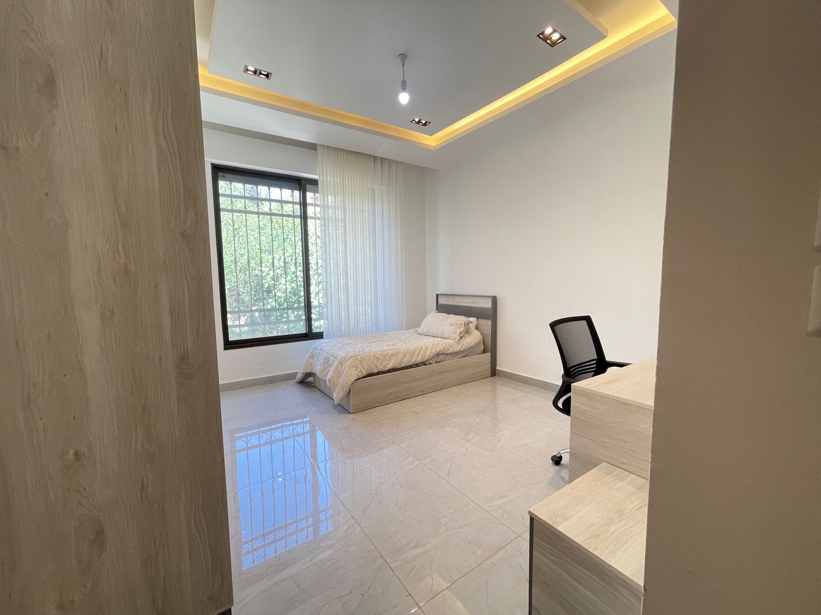 1271fe78-0a8b-4cdc-8bf9-2278e3b9f1cf Small bedroom with single bed, desk, and office chair; large window with bars and sheer curtain; glossy tiled floor and recessed ceiling lighting.