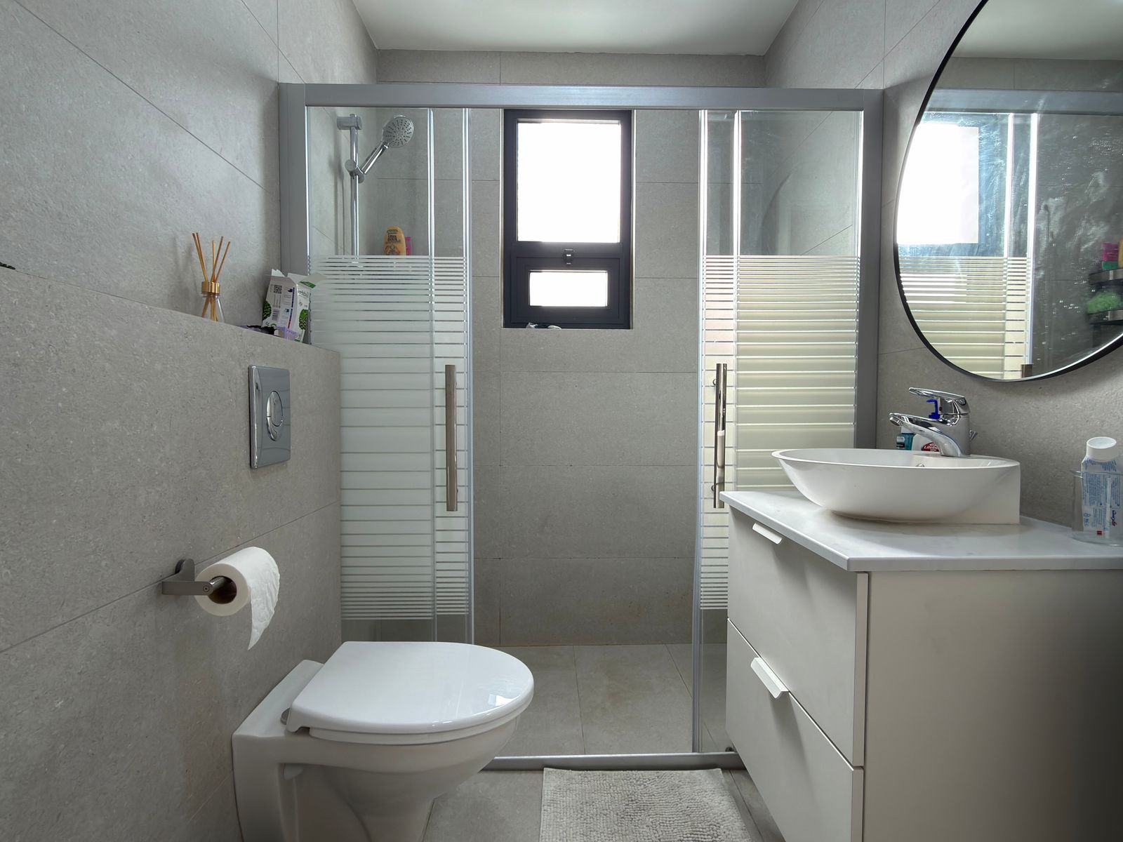 127be64c-1766-48da-ac4a-19af07864a88 Modern gray bathroom with hanging toilet, wall-mounted flush, a vanity with a round vessel sink and chrome faucet, glass shower doors, and a small window above.