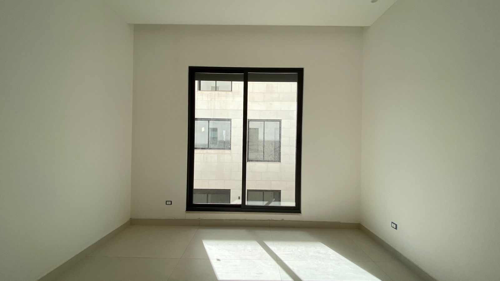 13251257-aa2a-41ea-ba5b-6f1c44edcb37 Empty white room with a large dark-framed window; sunlight casts a rectangular patch on the tiled floor, view of a beige building exterior outside.